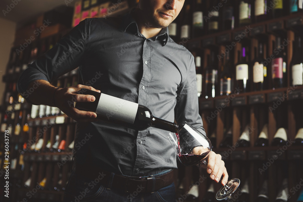 Close up shot of sommelier pouring red wine from bottle in glass on underground cellar background