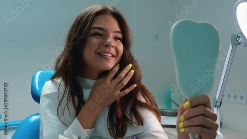 young beautiful brunette woman patient looks pleased with result watching in the mirror touching her teeth smiling while sitting in the dentist's office