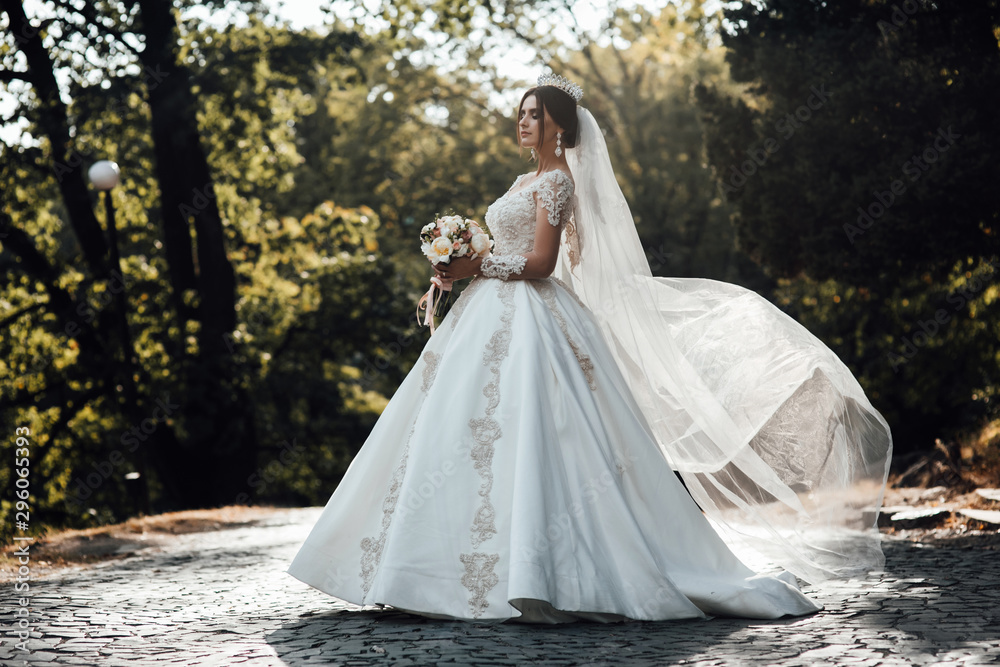 The bride in beautiful dress and with a bouquet in her hands for a walk. The wind waves the veil. Tender bride in wedding dress. Bouquet in bride's hands. Bride with hair style and make up The bride in beautiful dress and with a bouquet in her hands for a walk. The wind waves the veil. Tender bride in wedding dress. Bouquet in bride's hands. Bride with hair style and make up