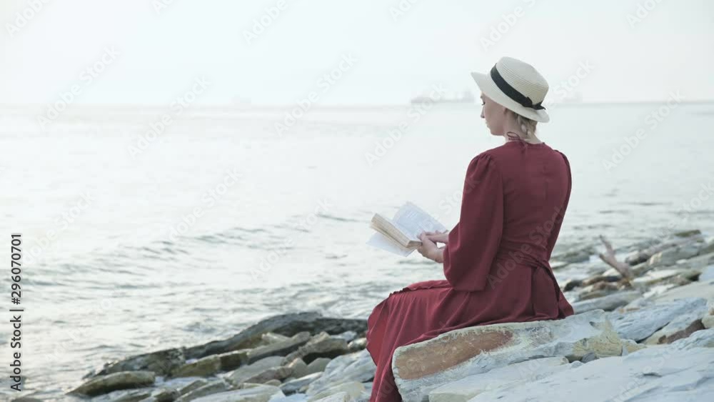 Back view An attractive Caucasian girl in a red bright dress and a straw hat sits on a large stone by the sea at sunset and reads an interesting book on the sea horizon and beautiful clouds. Back view An attractive Caucasian girl in a red bright dress and a straw hat sits on a large stone by the sea at sunset and reads an interesting book on the sea horizon and beautiful clouds.