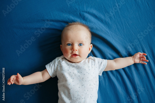 Portrait of cute adorable smiling white Caucasian baby girl boy with blue eyes four months old lying on bed in bedroom looking at camera. View from top above. Happy childhood lifestyle.