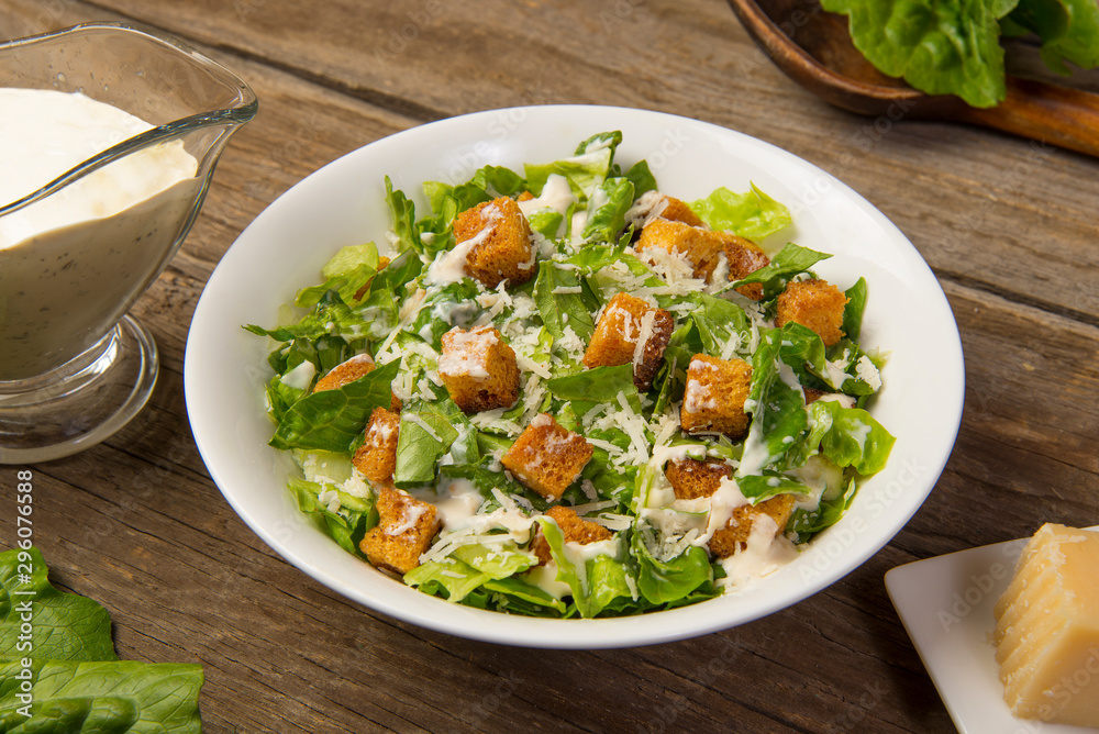 Caesar salad in a white bowl on an old rustic wooden table with leaves with a sauce boat and parmesan. Selected focus. Caesar salad in a white bowl on an old rustic wooden table with leaves with a sauce boat and parmesan. Selected focus.