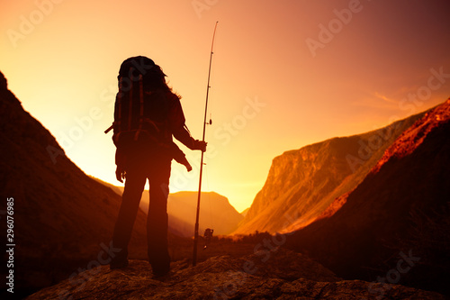 Woman with backpack stands on the rock with the fishing rod and enjoys sunrise valley view