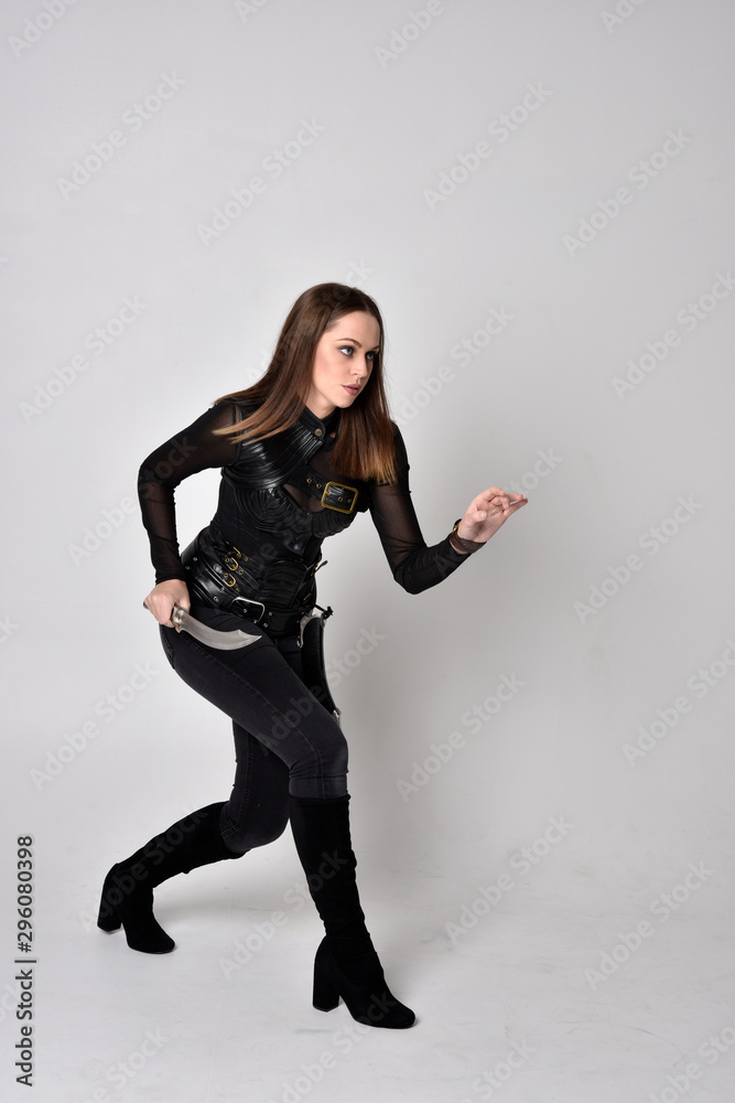 full length portrait of a pretty brunette woman wearing black leather fantasy costume. Standing pose, holding a dagger on a studio background. full length portrait of a pretty brunette woman wearing black leather fantasy costume. Standing pose, holding a dagger on a studio background.