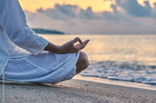 Woman in white clothes and a turban sits on a sandy beach at dawn and practices yoga
