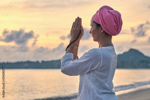 Woman in white clothes and a turban sits on a sandy beach at dawn and practices yoga