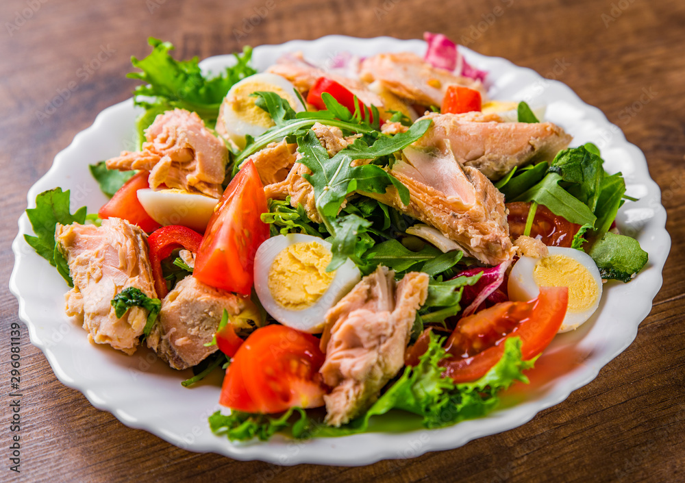 Fresh salad with fish, arugula, eggs,red pepper, lettuce, fresh sald leaves and tomato on a white plate on wooden table background Fresh salad with fish, arugula, eggs,red pepper, lettuce, fresh sald leaves and tomato on a white plate on wooden table background
