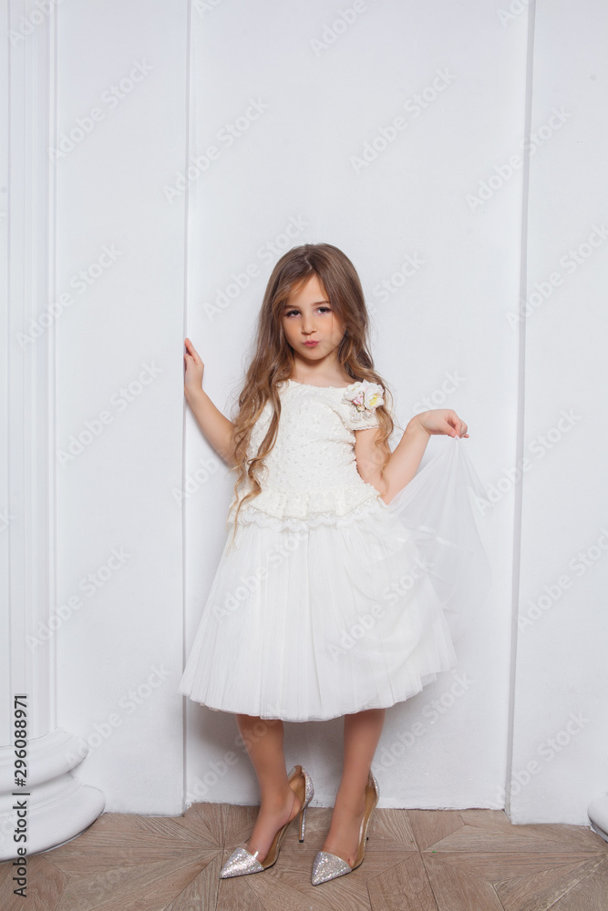 Little princess excited girl in fashion white dress wearing big mothers sparkle heels shoes on white background. Free space for text mockup Little princess excited girl in fashion white dress wearing big mothers sparkle heels shoes on white background. Free space for text mockup