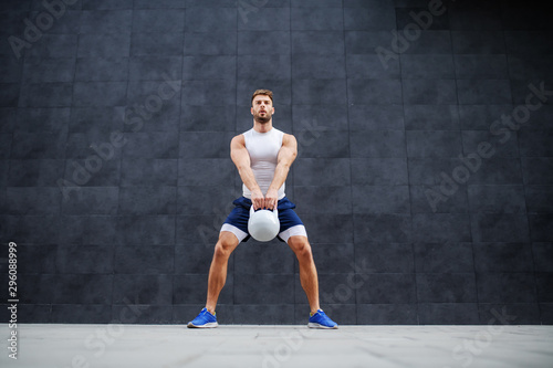 Strong muscular handsome Caucasian man in shorts and t-shirt standing outdoors and swinging kettle bell. In background is gray wall. © dusanpetkovic1