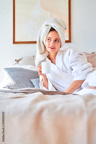 Photo of woman in white bathrobe with mug of tea in her hands lying on bed.
