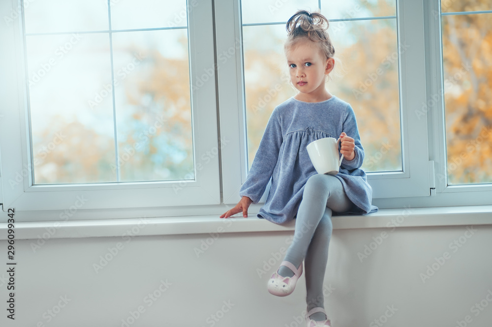 Cute cheerful little girl sitting by the big window with a cup of hot drink. Kids family house design expression. Cute cheerful little girl sitting by the big window with a cup of hot drink. Kids family house design expression.