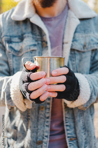 Man in mittens keeps warm holding a cup of hot drink. Chilly morning outdoors scene: human hands with a tin cup of hot beverage