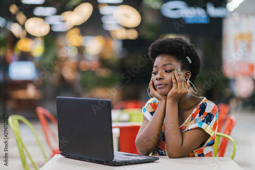 Young african american girl sitting at cafe table with laptop and looking at screen. Dark skinned freelancer concept. photo