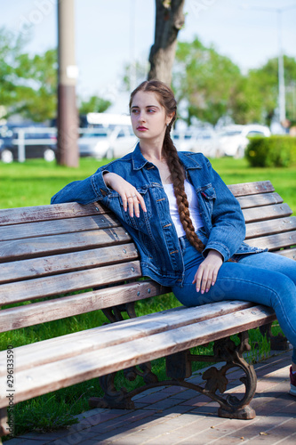 Beautiful girl in a denim jacket sits on a bench in a summer park