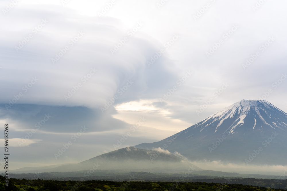 Mount Fuji before storm with cloudy hat on top , the light from sunset with dark clouds on overcast sky background, range of  dark forest trees, beautiful landmark in Fujinomiya Shizuoka Japan.