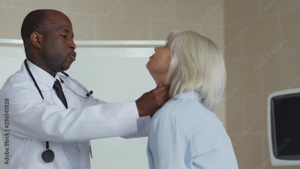 Medium shot side view of African American physician in white coat examining lymph nodes or tonsils of young female patient with blond hair during medical exam Medium shot side view of African American physician in white coat examining lymph nodes or tonsils of young female patient with blond hair during medical exam