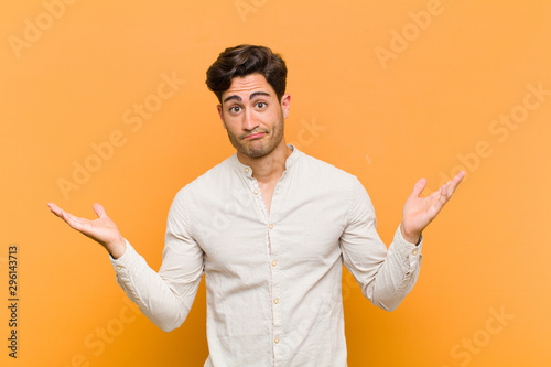 young handsome man feeling puzzled and confused, doubting, weighting or choosing different options with funny expression against orange background