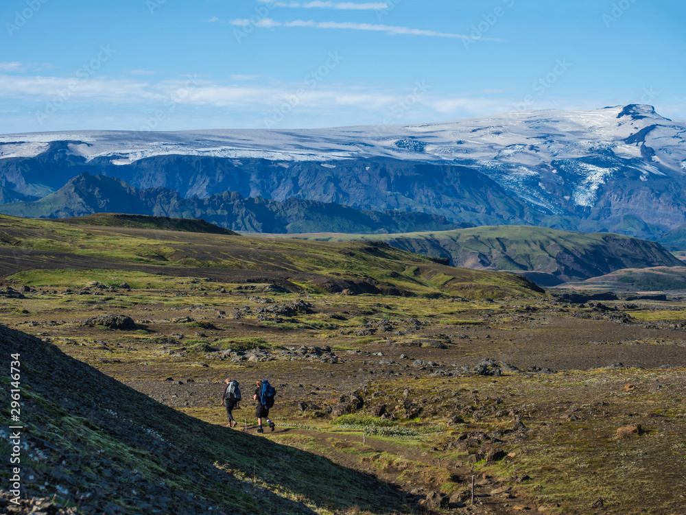 Couple of two young hikers at Laugavegur hiking trail with eyjafjallajokull volcano and glacier tongue, blue river stream and green hills. Fjallabak Nature Reserve, Iceland. Summer blue sky