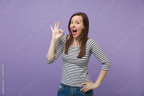 Shocked young brunette woman in casual striped clothes posing isolated on violet purple background studio portrait. People lifestyle concept. Mock up copy space. Keeping mouth open showing OK gesture.
