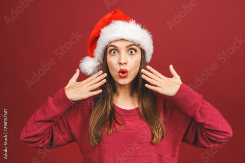 Attractive Woman With Santa Hat Looking Surprised And Amazed isolated against red background.