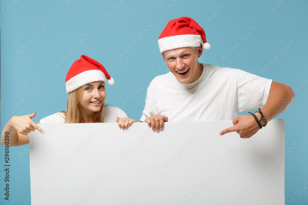 Smiling young Santa couple friends guy girl in Christmas hat isolated on blue background. Happy New Year 2020 celebration holiday concept. Mock up copy space. Holding big white empty blank billboard. Smiling young Santa couple friends guy girl in Christmas hat isolated on blue background. Happy New Year 2020 celebration holiday concept. Mock up copy space. Holding big white empty blank billboard.