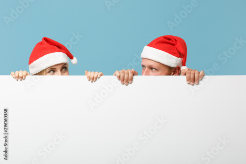 Pretty young Santa couple friends guy girl in Christmas hat isolated on blue background. Happy New Year 2020 celebration holiday concept. Mock up copy space. Holding big white empty blank billboard. © ViDi Studio