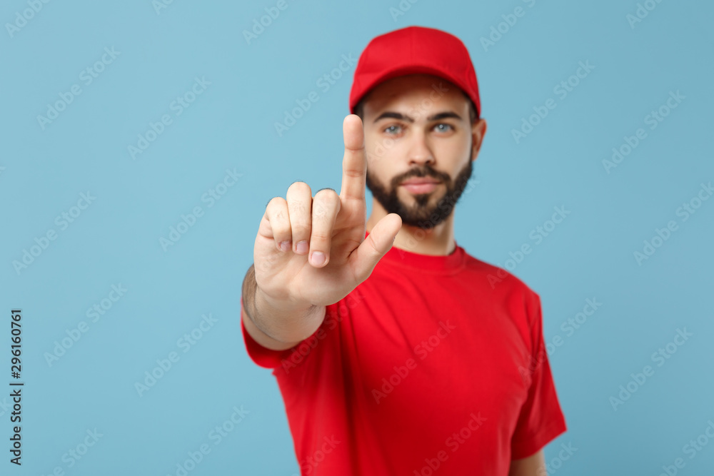 Delivery man in red uniform workwear isolated on blue wall background, studio portrait. Professional male employee in cap t-shirt print working as courier dealer. Service concept. Mock up copy space. Delivery man in red uniform workwear isolated on blue wall background, studio portrait. Professional male employee in cap t-shirt print working as courier dealer. Service concept. Mock up copy space.