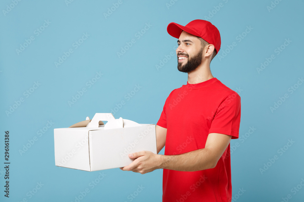 Delivery man in red uniform isolated on blue background, studio portrait. Male employee in cap t-shirt print courier hold cake dessert in unmarked cardboard box. Service concept. Mock up copy space.
