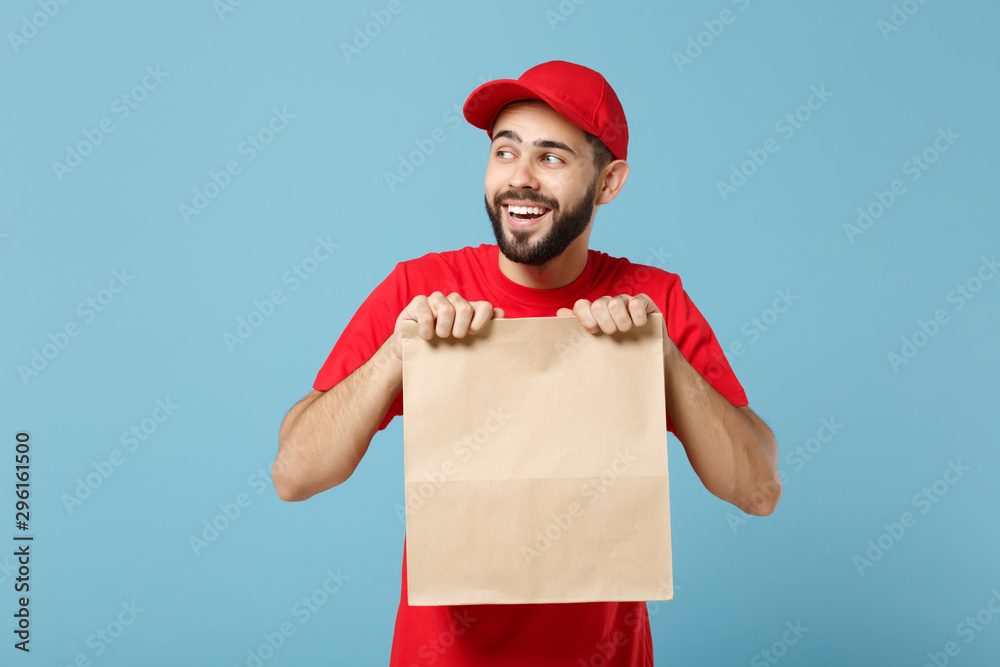 Delivery man in red uniform hold craft paper packet with food isolated on blue background, studio portrait. Male employee in cap t-shirt print working as courier. Service concept. Mock up copy space.