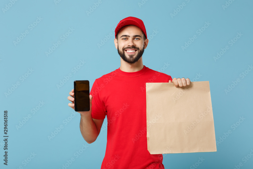 Delivery man in red uniform hold craft paper packet with food isolated on blue background, studio portrait. Male employee in cap t-shirt print working as courier. Service concept. Mock up copy space.