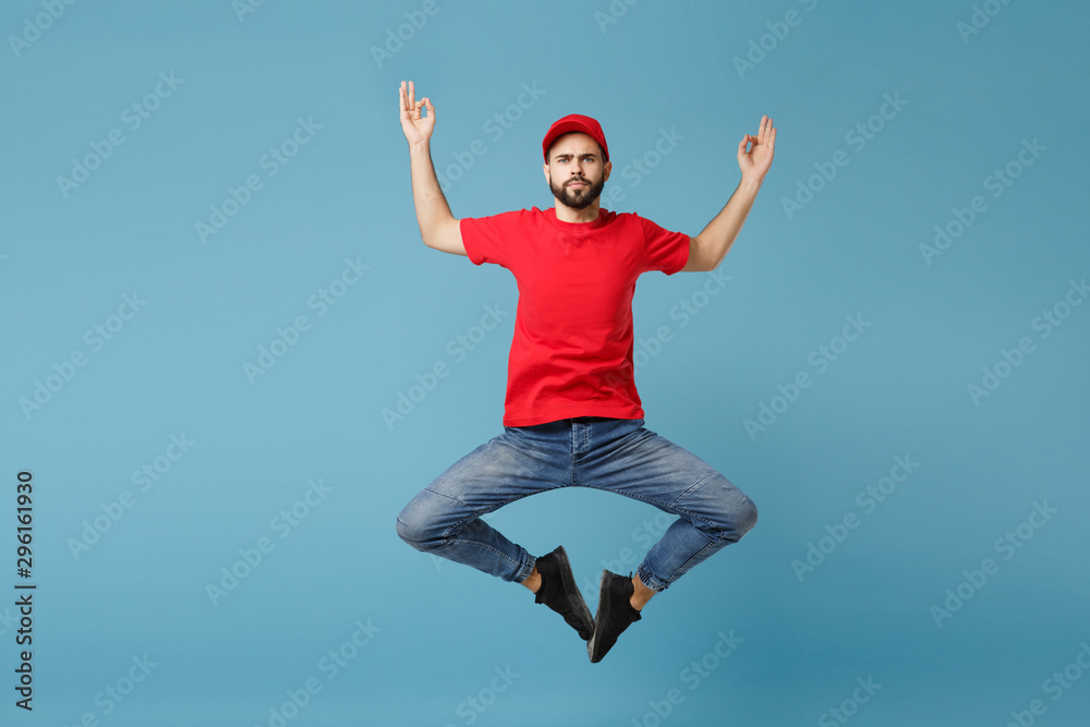 Delivery man in red uniform workwear isolated on blue wall background, studio portrait. Professional male employee in cap t-shirt print working as courier dealer. Service concept. Mock up copy space. Delivery man in red uniform workwear isolated on blue wall background, studio portrait. Professional male employee in cap t-shirt print working as courier dealer. Service concept. Mock up copy space.