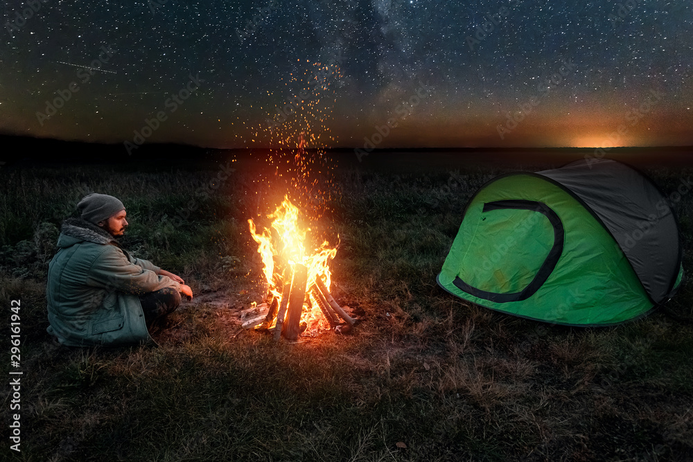 Camping man sits by the fire at night against the background of the starry sky. The concept of travel, tourism, camping. Camping man sits by the fire at night against the background of the starry sky. The concept of travel, tourism, camping.
