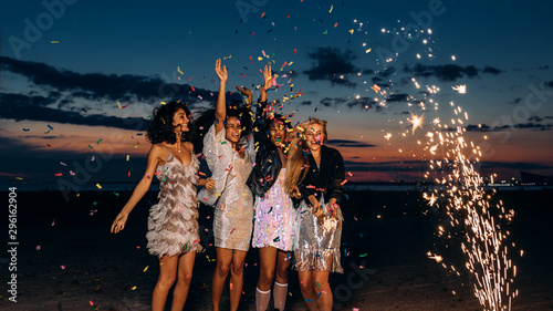 Group of four girlfriends dancing under confetti at sunset. Happy women celebrating with fireworks outdoors at evening. photo