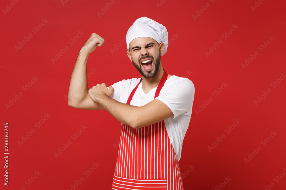 Strong young bearded male chef cook or baker man in striped apron white t-shirt toque chefs hat posing isolated on red background. Cooking food concept. Mock up copy space. Showing biceps, muscles. Strong young bearded male chef cook or baker man in striped apron white t-shirt toque chefs hat posing isolated on red background. Cooking food concept. Mock up copy space. Showing biceps, muscles.