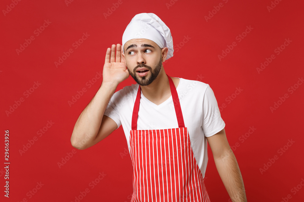 Curious bearded young male chef cook or baker man in striped apron white t-shirt toque chefs hat posing isolated on red background in studio. Cooking food concept. Mock up copy space. Try to hear you. Curious bearded young male chef cook or baker man in striped apron white t-shirt toque chefs hat posing isolated on red background in studio. Cooking food concept. Mock up copy space. Try to hear you.
