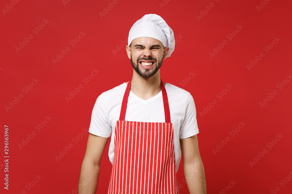 Angry irritated young bearded male chef cook baker man in striped apron white t-shirt toque chefs hat posing isolated on red wall background. Cooking food concept. Mock up copy space. Showing teeth. Angry irritated young bearded male chef cook baker man in striped apron white t-shirt toque chefs hat posing isolated on red wall background. Cooking food concept. Mock up copy space. Showing teeth.