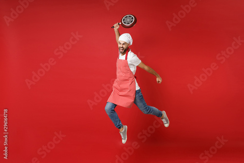 Crazy young bearded male chef cook or baker man in striped apron white t-shirt toque chefs hat posing isolated on red background. Cooking food concept. Mock up copy space. Jumping, holding frying pan.