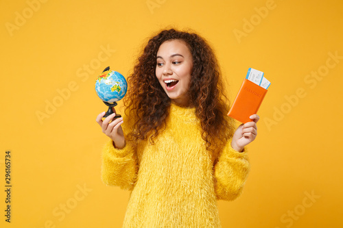 Excited young african american girl in fur sweater posing isolated on yellow orange wall background. People lifestyle concept. Mock up copy space. Holding world globe, passport, boarding pass tickets.