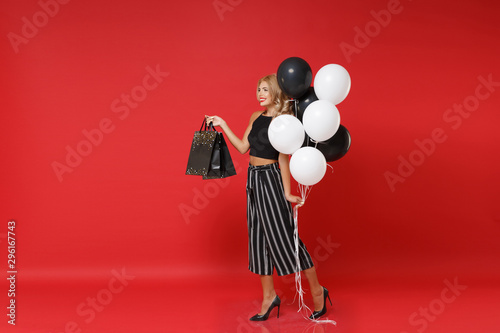 Smiling woman girl in black clothes posing isolated on red background. Shopping discount sale, holiday party concept. Mock up copy space. Celebrating, holding air balloons, package bag with purchases.