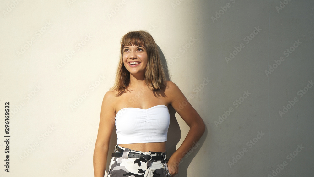 young blond woman leaning against a wall in a shaft of sunlight while smiling and looking away young blond woman leaning against a wall in a shaft of sunlight while smiling and looking away