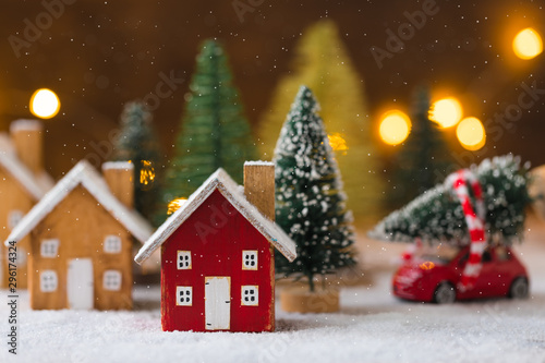 Miniature wooden houses and small red car with fir tree on the snow over blurred Christmas decoration background, toned