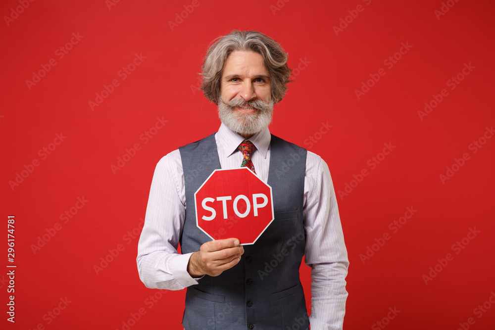 Smiling elderly gray-haired mustache bearded man in classic shirt vest and colorful tie isolated on red wall background. People lifestyle concept. Mock up copy space. Holding red sign with Stop title. Smiling elderly gray-haired mustache bearded man in classic shirt vest and colorful tie isolated on red wall background. People lifestyle concept. Mock up copy space. Holding red sign with Stop title.