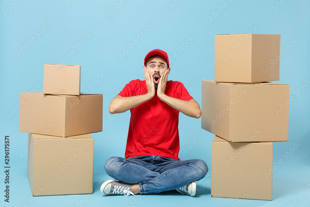 Delivery man in red uniform isolated on blue background, studio portrait. Male employee in cap t-shirt print working as courier dealer hold empty cardboard box. Service concept. Mock up copy space. Delivery man in red uniform isolated on blue background, studio portrait. Male employee in cap t-shirt print working as courier dealer hold empty cardboard box. Service concept. Mock up copy space.
