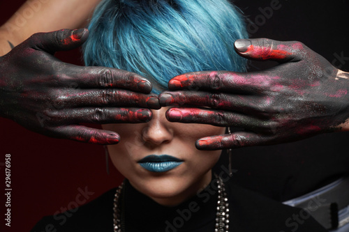 Eyes of beautiful girl with short blue fashionable hairstyle and blue lipstick covered by men's hands in black, pink and red paint on a dark background. See no evil concept. Close-up studio photo