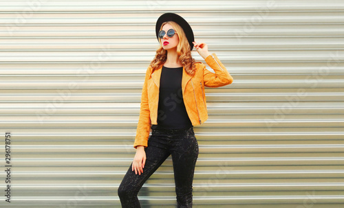 Stylish woman model posing wearing yellow jacket, black round hat on metal wall background