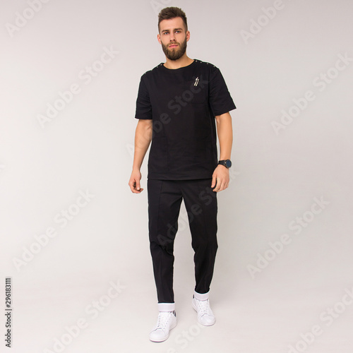 young stylish bearded nurse male in black medical costume is standing straight and smiling cute on the white wall background. medical fashion concept. free space