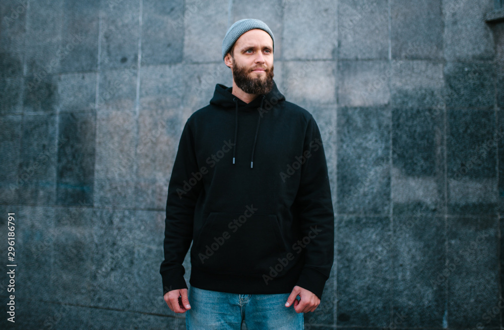 City portrait of handsome hipster guy with beard wearing black blank hoodie or sweatshirt and hat with space for your logo or design. Mockup for print City portrait of handsome hipster guy with beard wearing black blank hoodie or sweatshirt and hat with space for your logo or design. Mockup for print