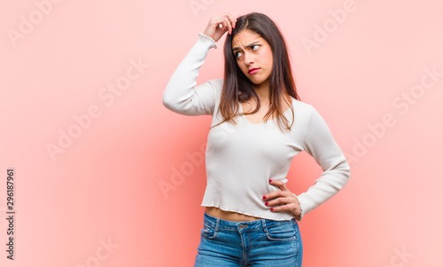 Bild auf Leinwand young  pretty woman feeling puzzled and confused, scratching head and looking to the side against pink background