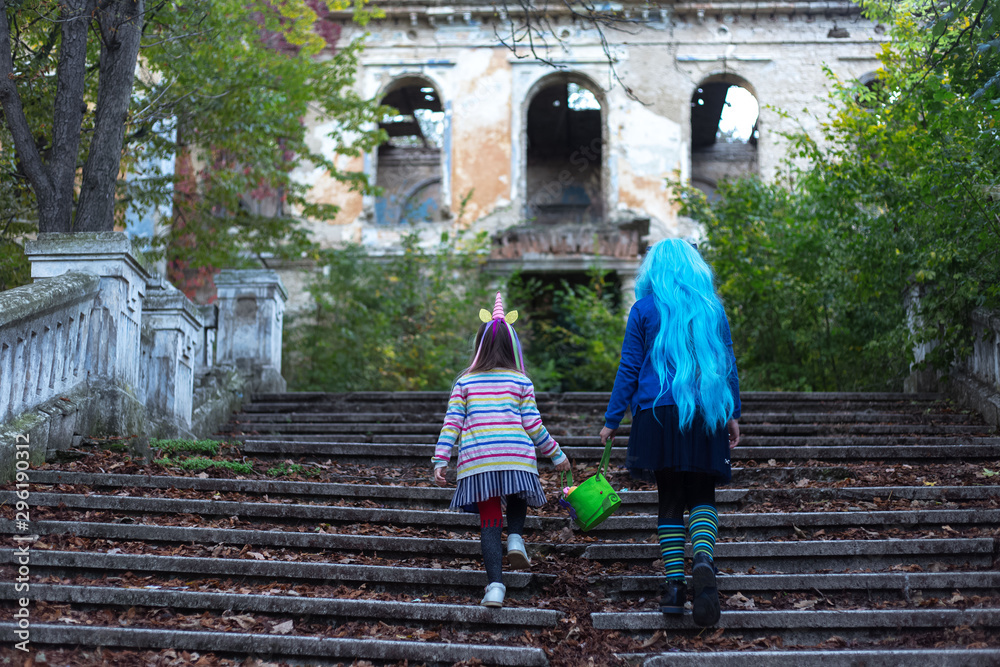 Back view of two kids sister in halloween costume of unicorn, with halloween bucket, walking up stairs to old abandoned house.