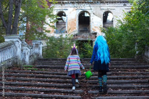 Back view of two kids sister in halloween costume of unicorn, with halloween bucket, walking up stairs to old abandoned house. © Lalandrew